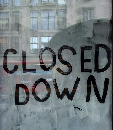 The words "closed down" written on a dirty window with a reflection of a building across the street.