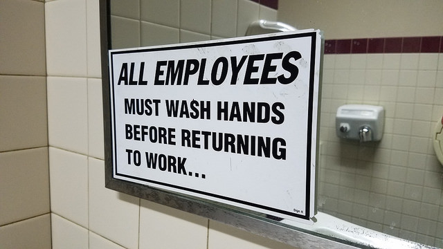 "All Employees Must Wash Hands Before Returning to Work" sign on mirror with reflection of hand dryer in background