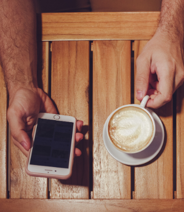 SCS, image of a person with a smart phone in one hand and a coffee cup in the other, sitting at a table 